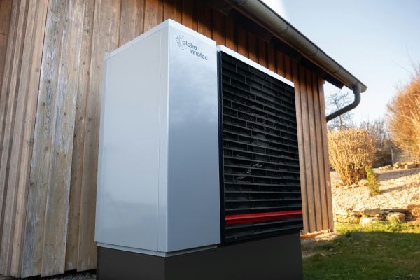 Moderne Außeneinheit einer Wärmepumpe von Alpha Innotec vor einer Holzhauswand im Gartenbereich.
