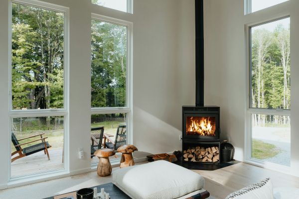 Moderner Kaminofen mit sichtbarem Holzlager in hellem Wohnzimmer mit großen Fenstern und Blick ins Grüne.